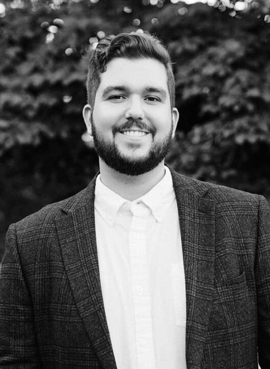 Black and white headshot of Metrospect Media owner Jason. He is wearing a white shirt with a dark jacket and standing in front of a leafy background looking straight at the camera. 