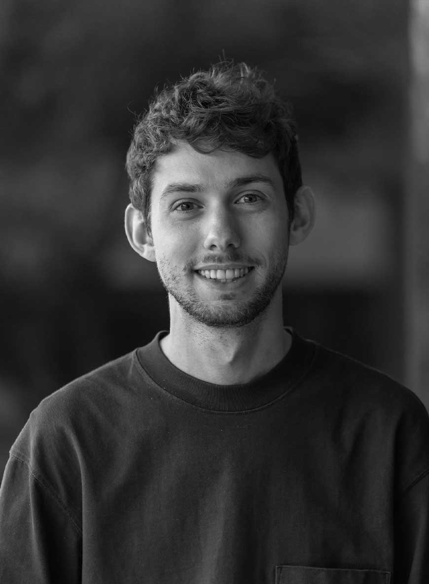 Black and white headshot of lead editor Jordan. He is wearing a dark colored shirt and smiling at the camera against a blurred background.