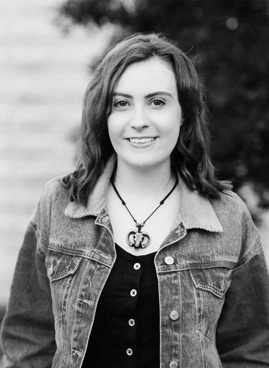 Black and white headshot of editor Micah. She has dark hair and is wearing a necklace and a denim jacket while smiling into the camera. 