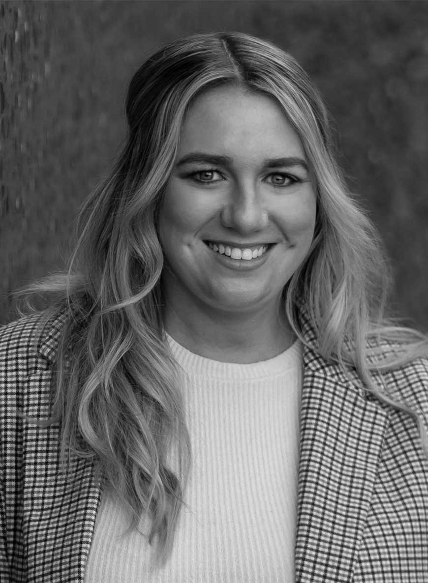 Black and white headshot of co-owner Molly. She is wearing a white shirt with a patterned blazer and is standing against a textured wall. 