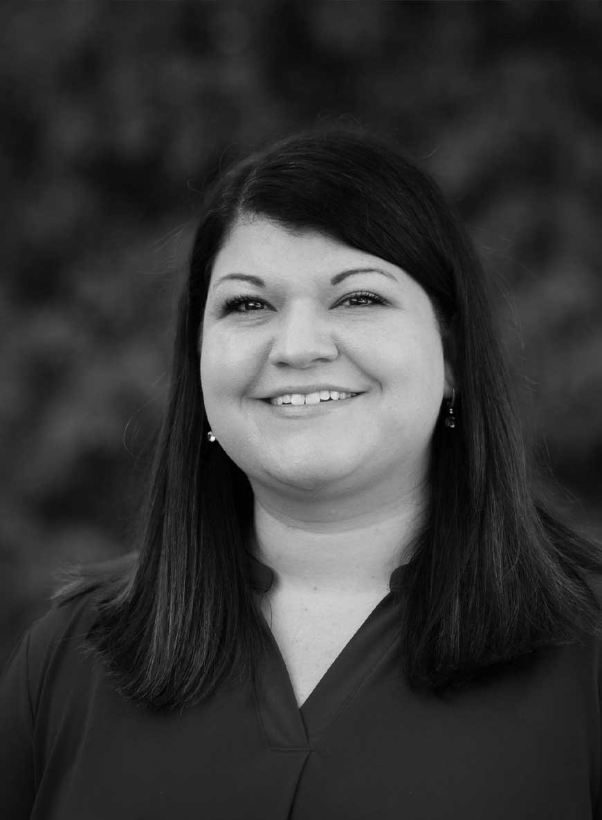 Black and white headshot of director of operations Sarah. She has dark hair and is smiling widely at the camera.