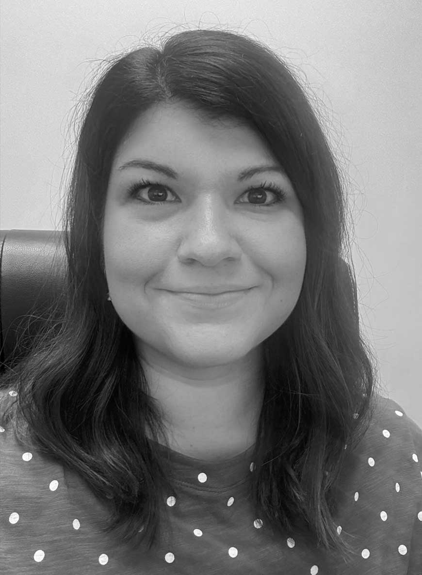 Black and white headshot of director of operations Sarah. She has dark hair and is smiling widely at the camera.